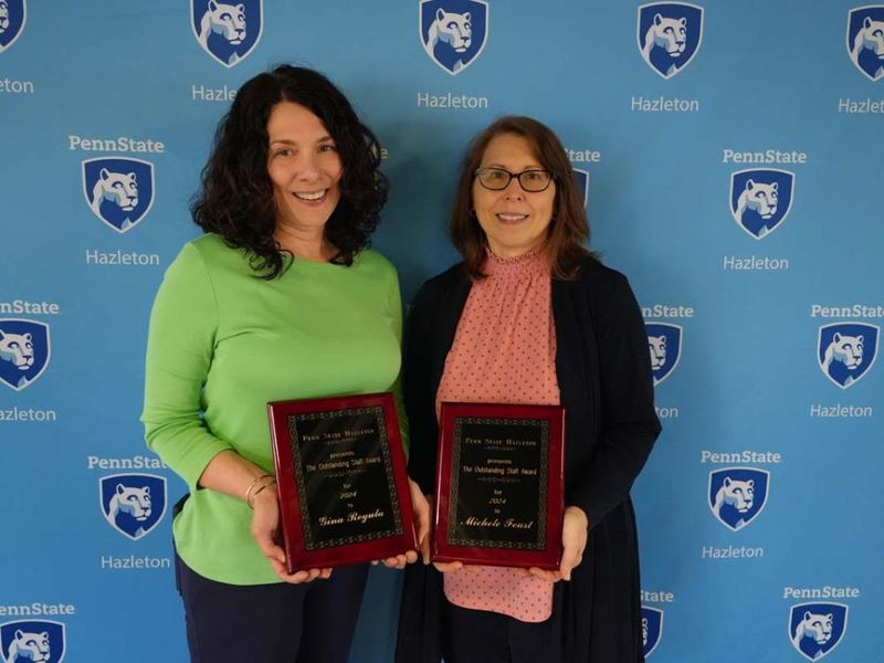 Two women holding plaques and standing in front of light blue Penn State Hazleton backdrop.