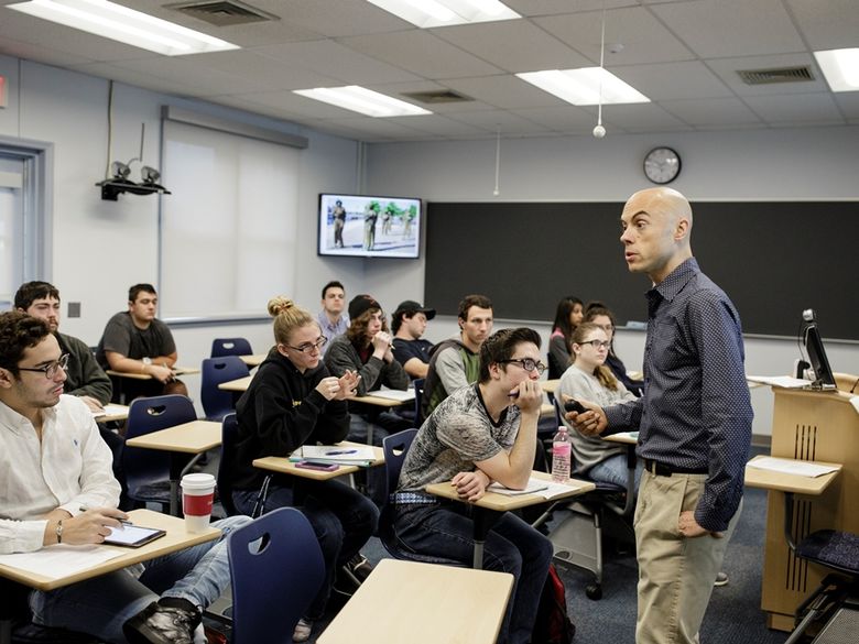 Professor pointing to white board on wall in classroom filled with students.